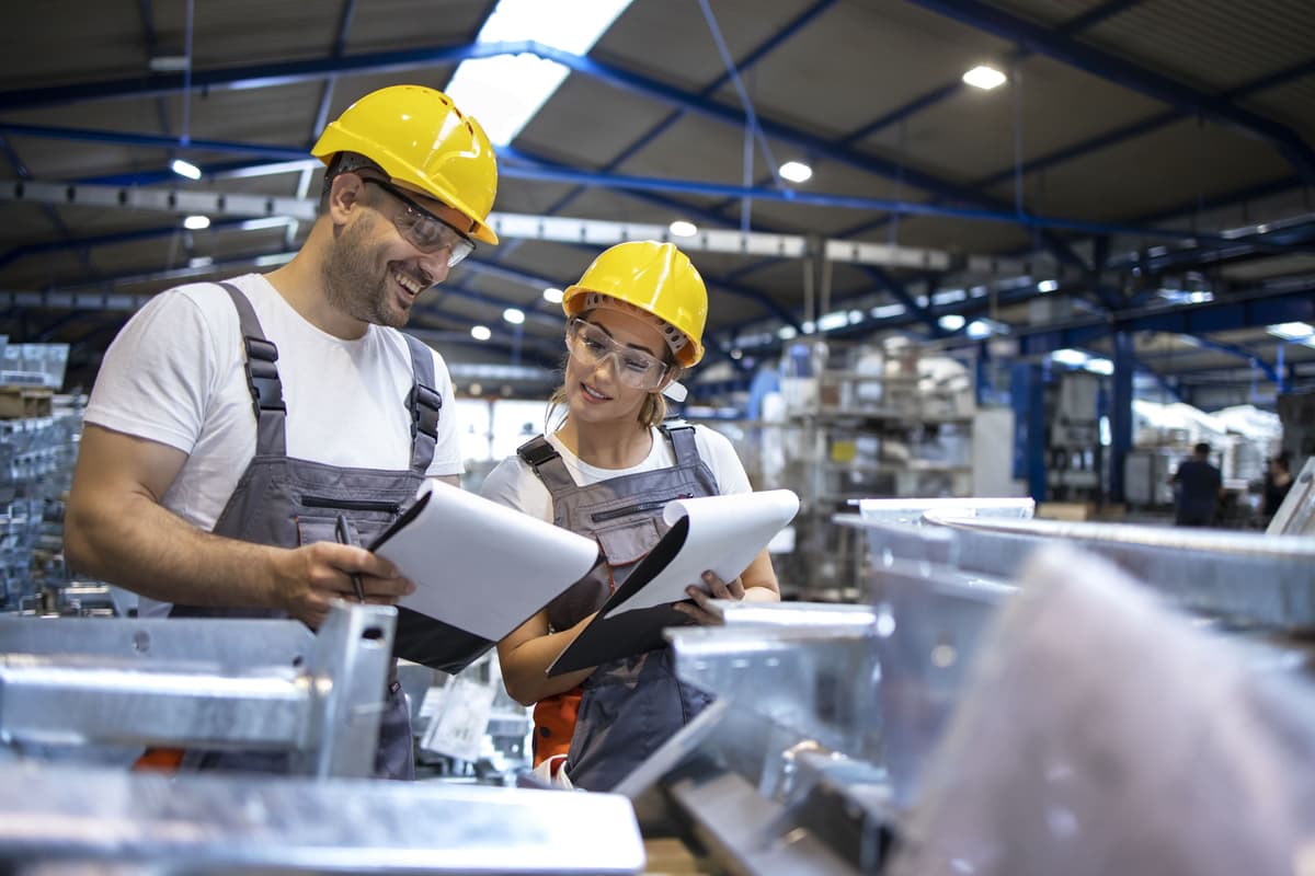 Dois funcionários de fábrica com capacete e óculos de proteção conferindo uma peça