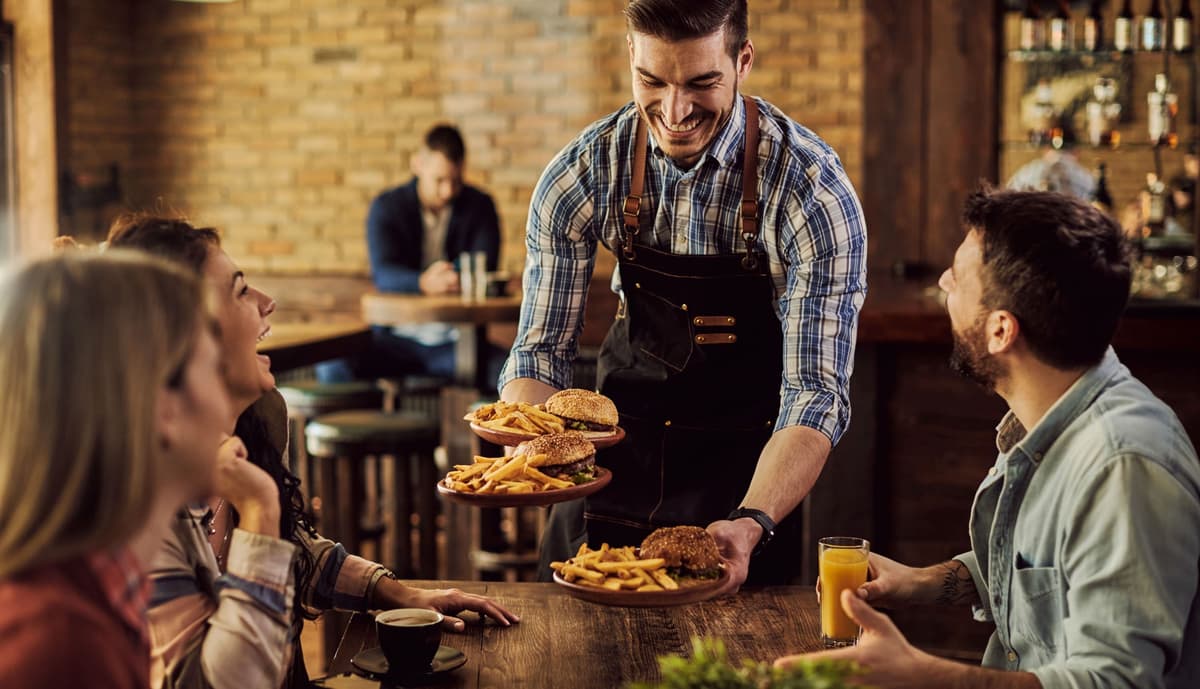 Garçom sorridente servindo pratos de hambúrguer e batata frita para clientes no balcão
